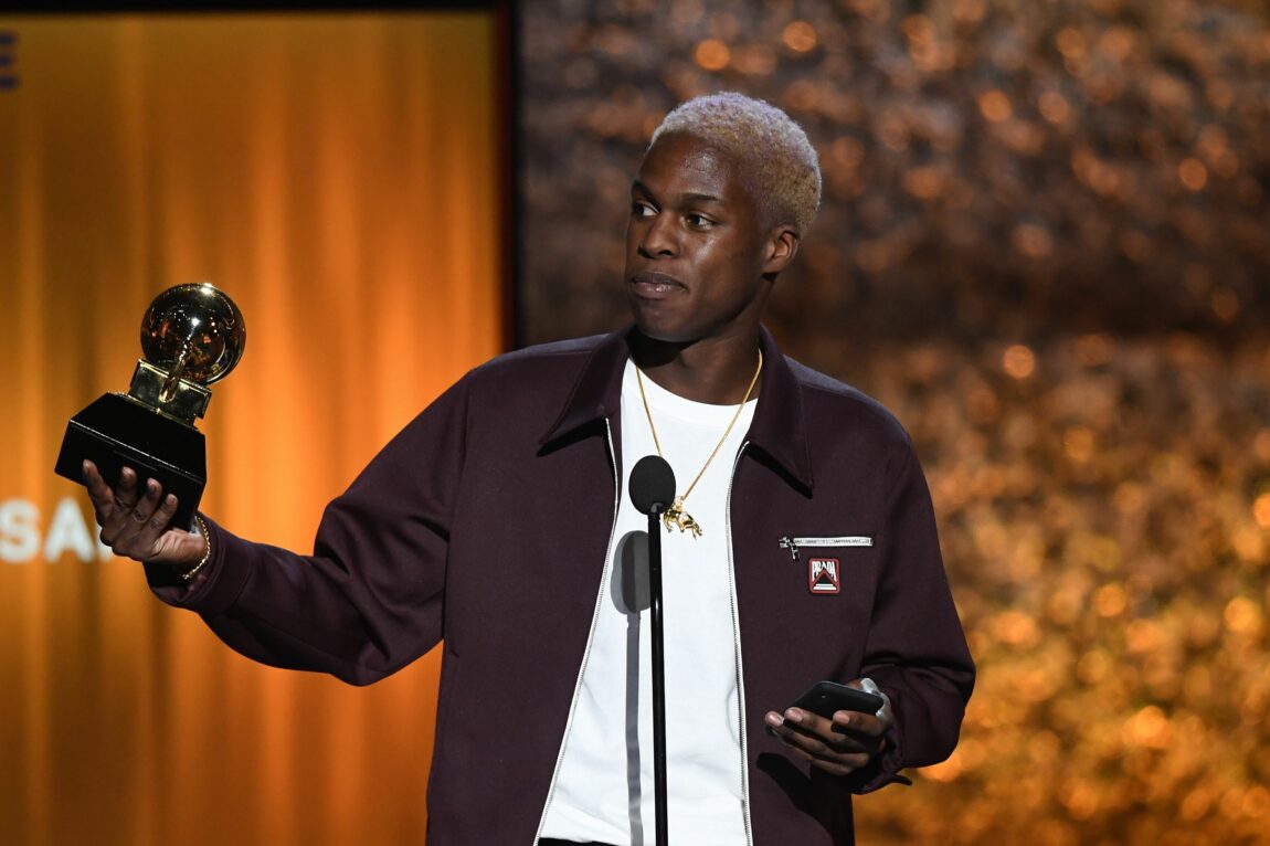 Daniel Caesar har på seg en svart jakke og en hvit t-skjorte. Han har farget håret blondt og holder opp en grammy.