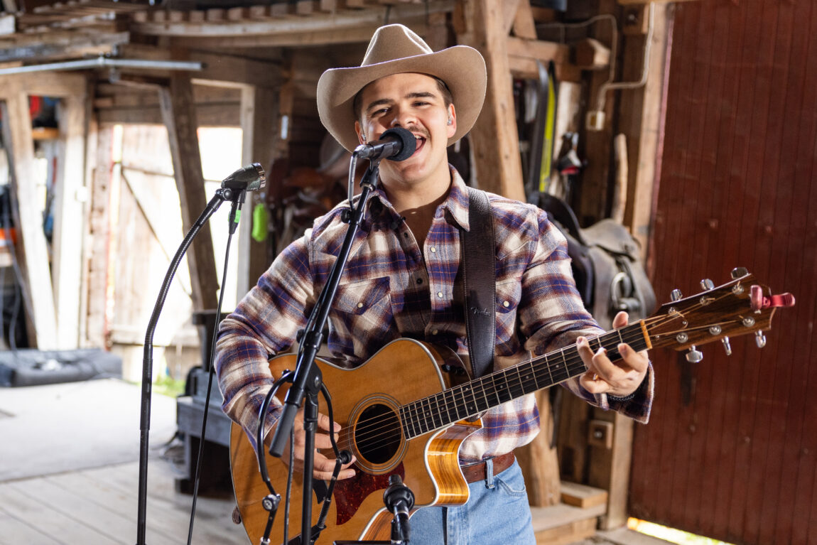 Tobias Sten spiller gitar og synger i mikrofonen. Har på seg en rutete skjorte, cowboyhatt og skjorta ned i buksa.