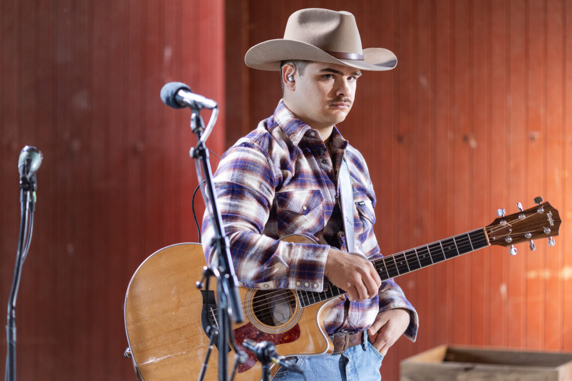 Tobias Sten spiller gitar og synger i mikrofonen. Har på seg en rutete skjorte, cowboyhatt og skjorta ned i buksa.