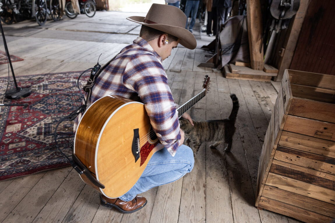 Tobias Sten har på seg en rutete skjorte, cowboyhatt og skjorta ned i buksa. Her klapper han en katt