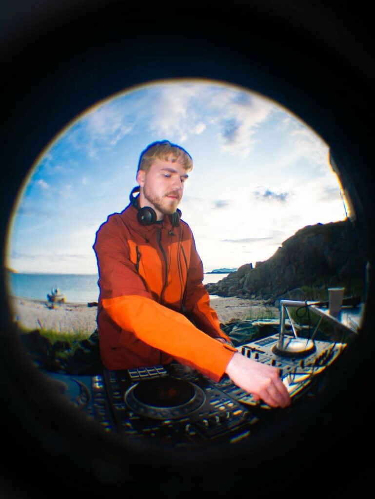 Vebjørn er DJ utendørs på en strand. Han har på seg en rød allværsjakke og spinner plate.