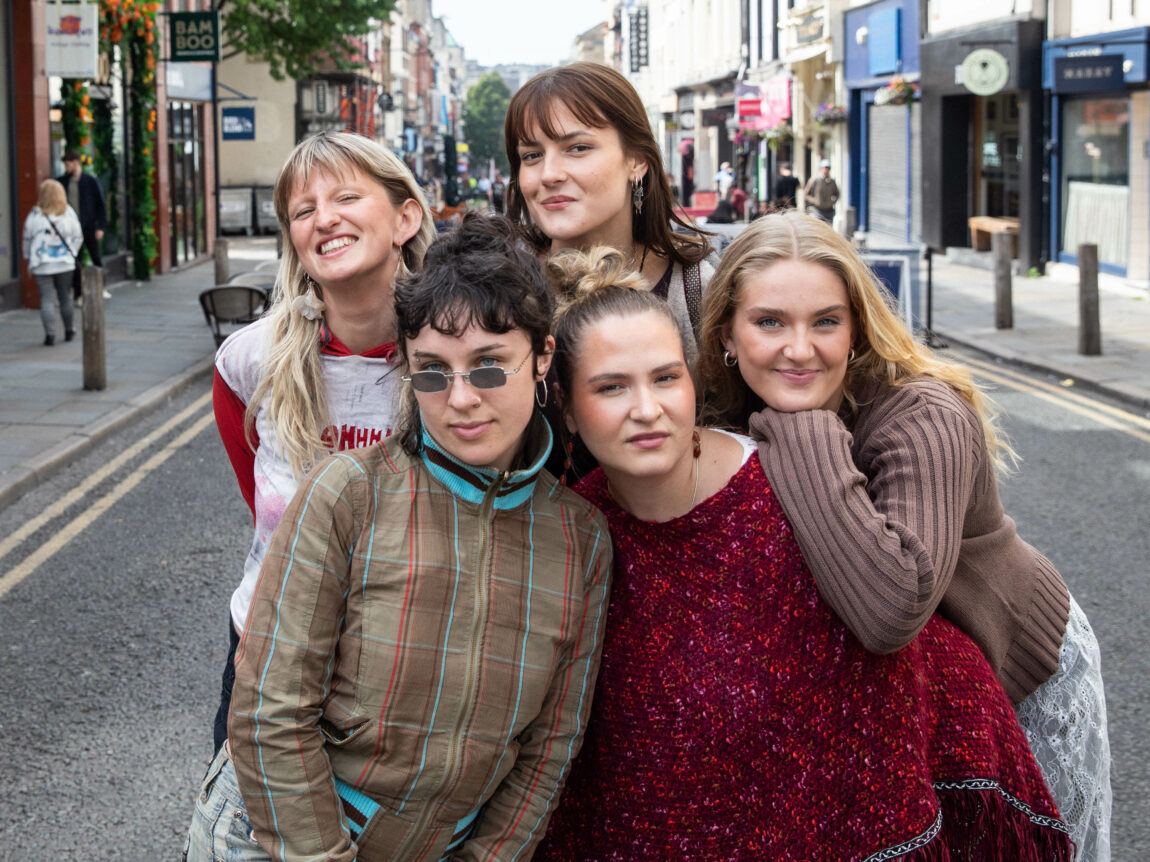 Girl Group står intill hverandre og lener seg på hverandre og ser tøysete inn i kamera. Bak dem er det et yrende gateliv i Liverpool 