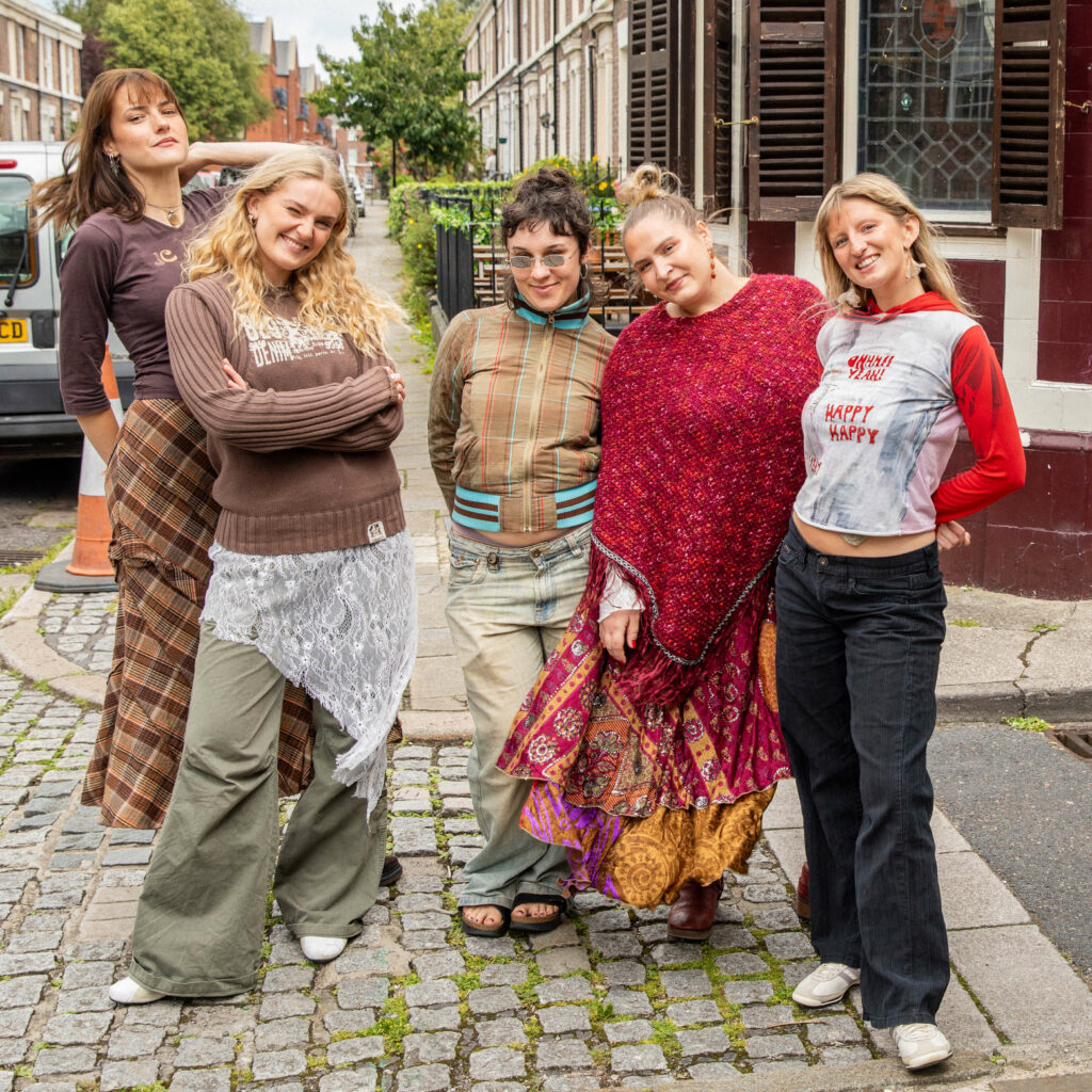 Girl Group poserer og smiler midt i gata i Liverpool. Bak dem er det fortau og en park.