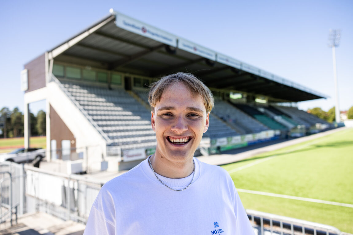 August har på seg en hvit t-skjorte og smiler enormt inn i kamera. Bak han er fotballbanen i Hønefoss.