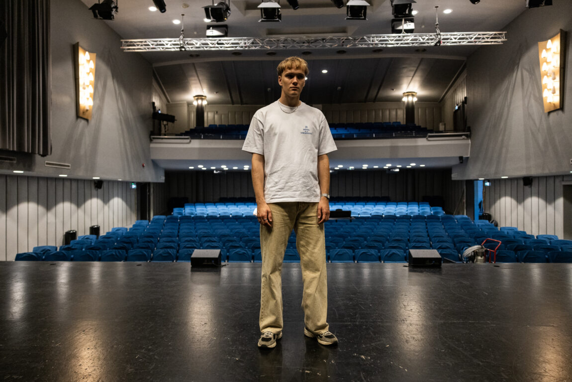 August har på seg en hvit t-skjorte. Han står på scenen i et teater. Bak han er det seter som er blå.