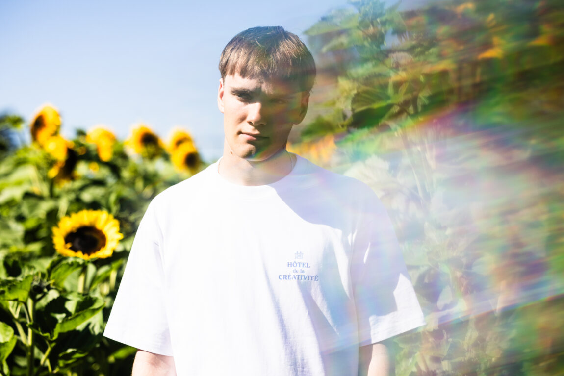 August har på seg en hvit t-skjorte. Han står midt i en blomstereng. Og ser seriøst inn i kamera.