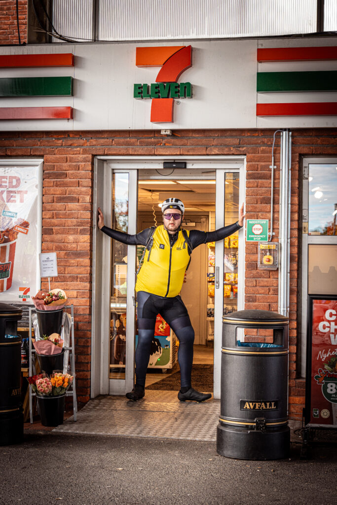 Jonas poserer mellom dørene på 7/11 et sted mellom Oslo og Kongsberg, han har på seg sykkelshorts med tights under, sykkelvest, hjelm og solbriller.