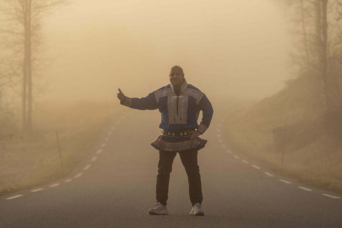 Fred Buljo står i blå og rød kofte langs veien og holder ut tommelen i en haik