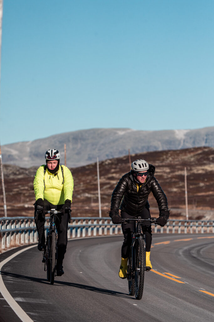 Carl Fredrik sykler ved siden av Jonas i svart boblejakke, Jonas har på seg refleksgul sykkeljakke. Det er sol og fjell i bakgrunnen.