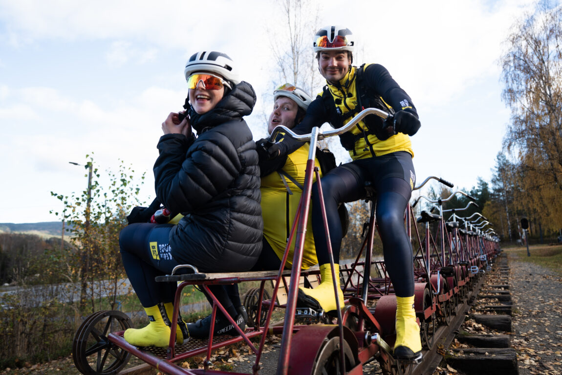 Anna med solbriller og hjelm og boblejakke sitter frmest på Dresinen, Sindre sitter med henda på styret, Jonas lener seg mot Sindre.