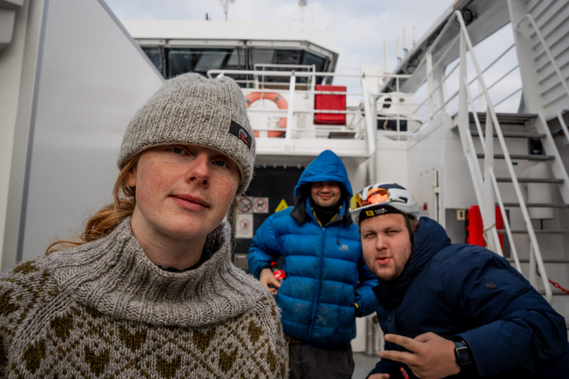 Anna kikker med et uttrykksløst fjes inn i kamera med grå lue og grå strikkagenser, JOnas poserer med peace-tegn i bakgrunnen og sindre smiler i bakgrunnen under en blå hette.