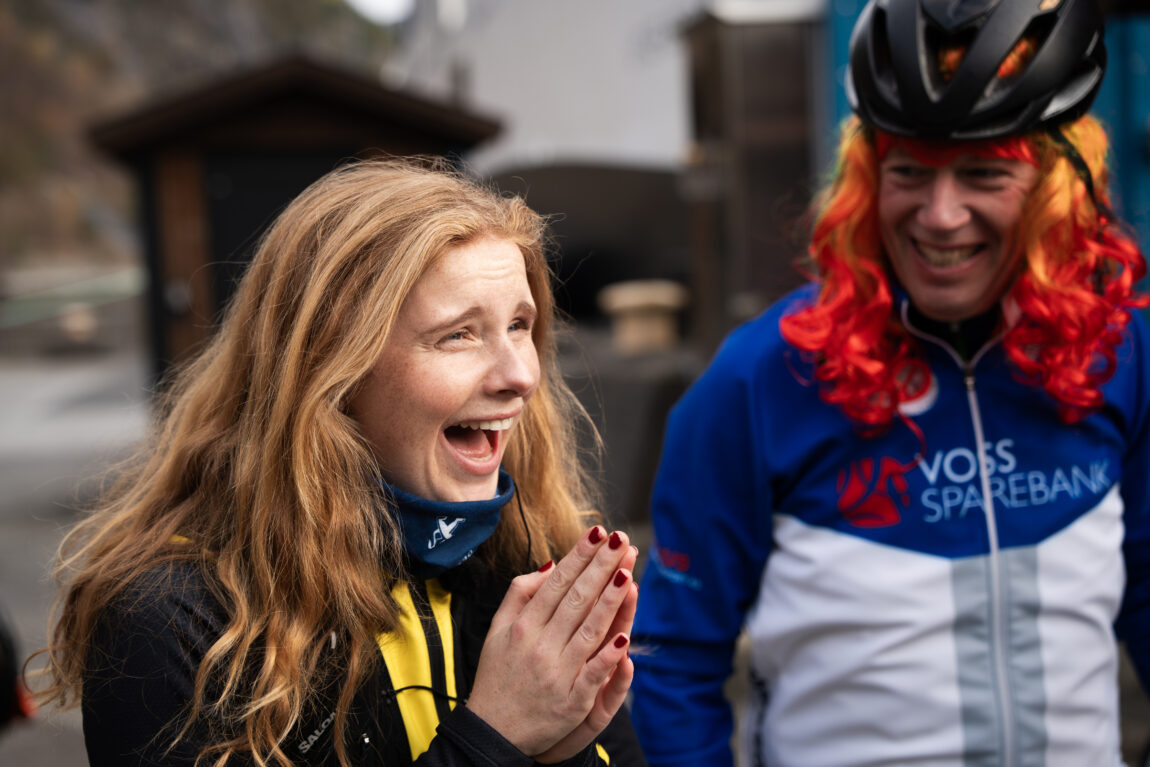 Anna ler og holder hendene samlet mens hun ser på gjengen med rødhårete parykker. I bakgrunnen ser man en glisende sykkelmann med rød parykk under hjelmen.