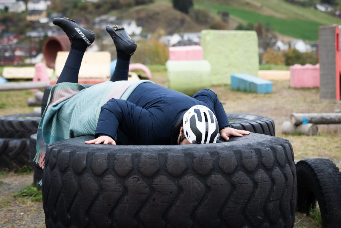 Jonas stuper nedi et bildekk med hodet først og med sykkelsko, sykkelhjelm på og et ullpledd rundt seg.