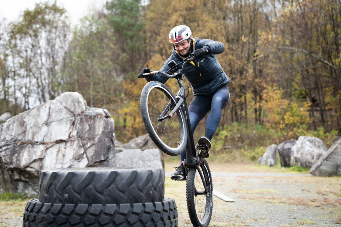 Sindre steiler nesten opp på to stabla bildekk på en trailsykkel, iført sykkeltights, hvit hjelm og tynn boblejakke.